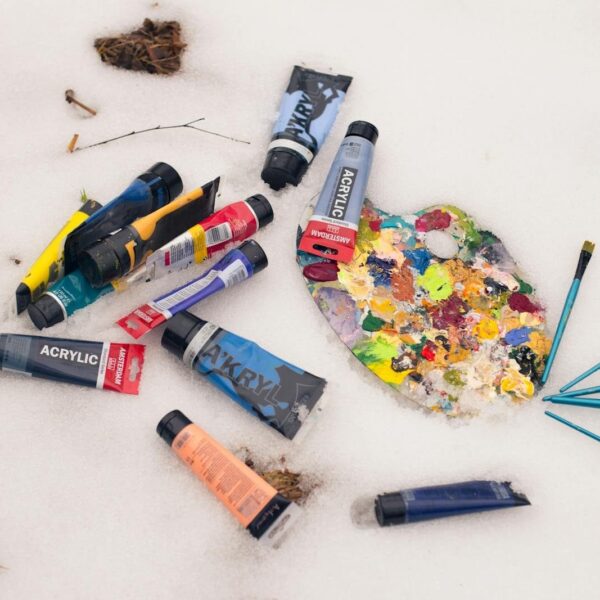 Colorful acrylic paint tubes and palette with brushes lying on a snow surface, artistic winter theme.