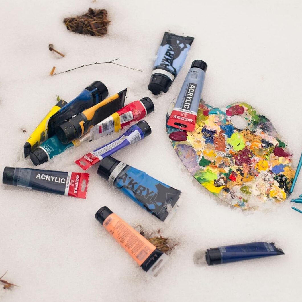 Colorful acrylic paint tubes and palette with brushes lying on a snow surface, artistic winter theme.