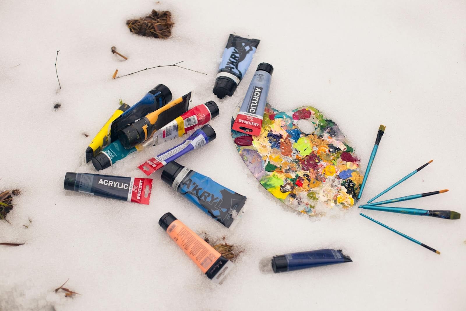 Colorful acrylic paint tubes and palette with brushes lying on a snow surface, artistic winter theme.